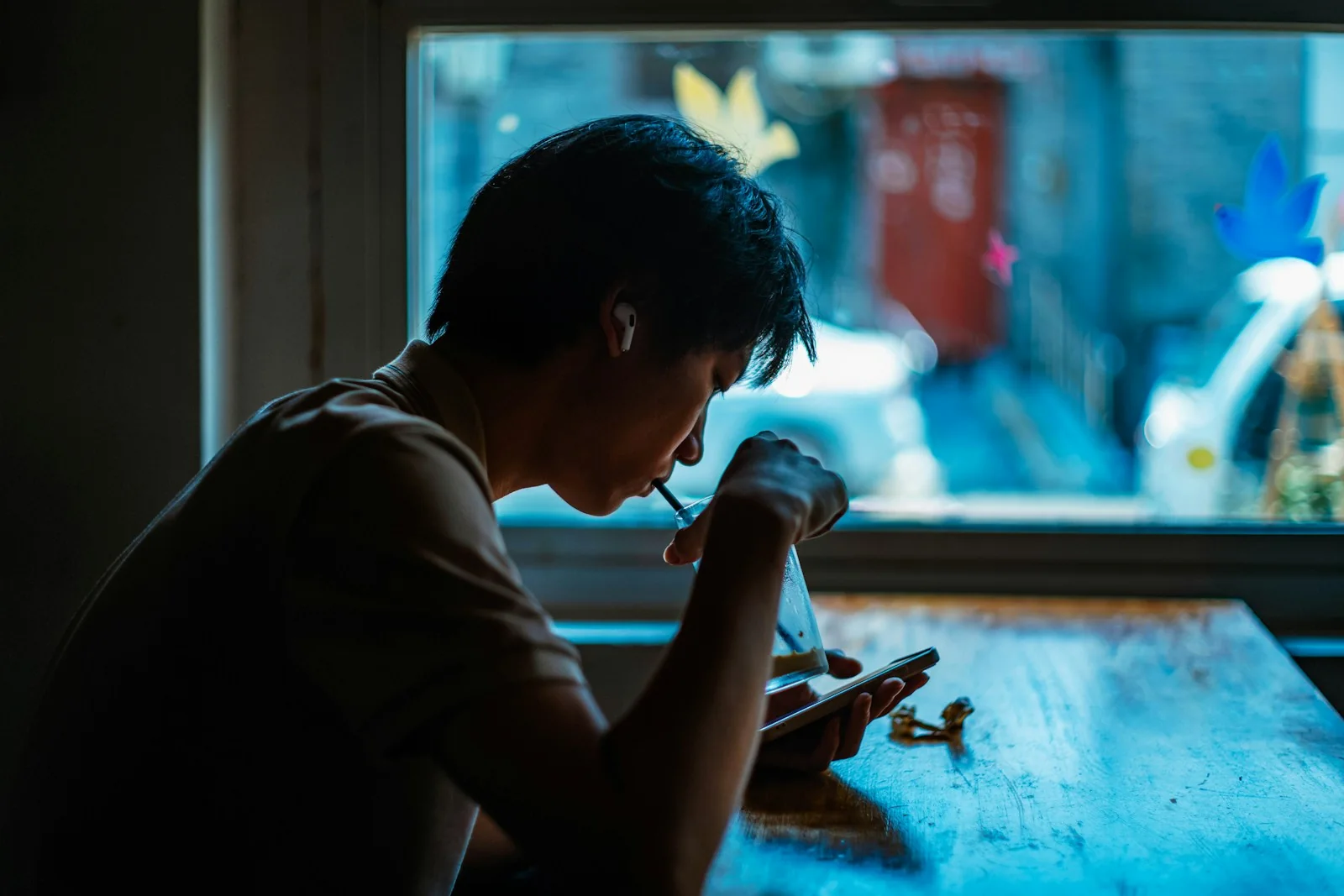 Un chico come solo junto a una ventana, con la cuchara en la boca y los ojos en el móvil