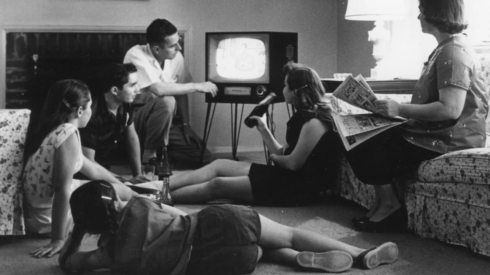 Una familia de 1958 ve la televisión en el salón: los hijos sentados en la alfombra, el padre ajusta el aparato y la madre lee el periódico en el sofá